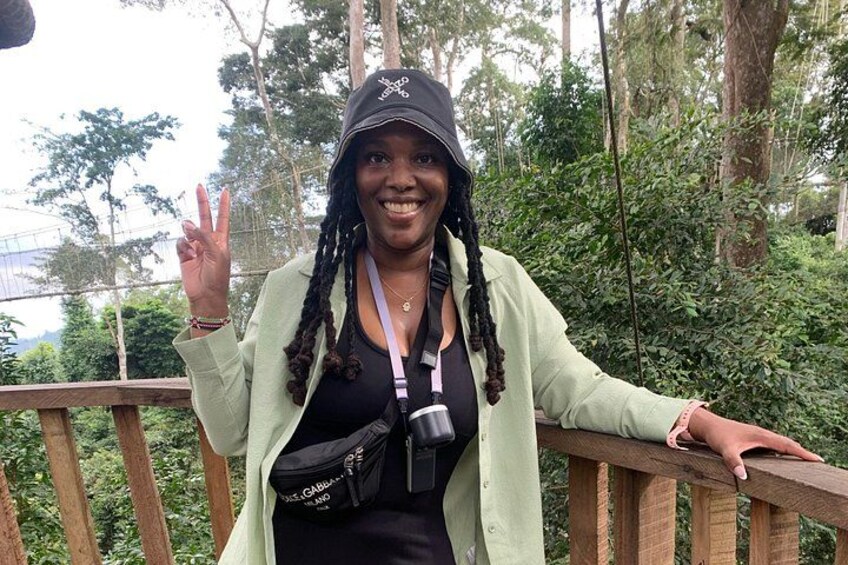 A person wearing a light green jacket and a black cap, standing on a wooden deck with greenery in the background. 4