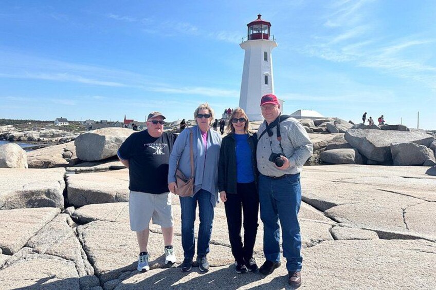 A group of people standing in front of a lighthouse, with large rocks and a clear sky in the background. 5