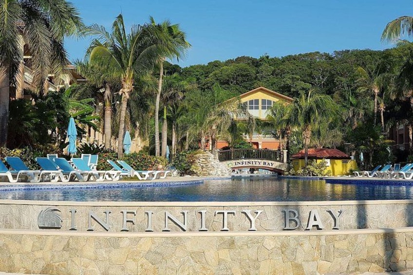 Ett poolområde med solstolar, en skylt med texten ”Infinity Bay”, och en byggnad omgiven av palmer. 5