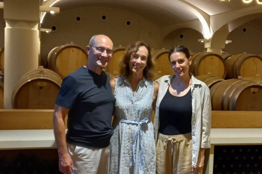 Three people standing in a wine cellar with wooden barrels in the background. 3