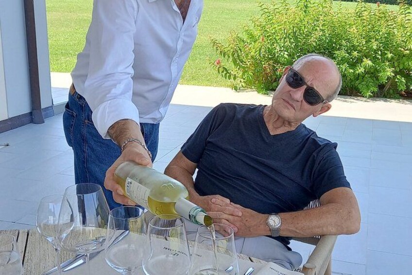 An outdoor setting with a table set for a meal, a person pouring wine into a glass, and a person seated at the table. 2