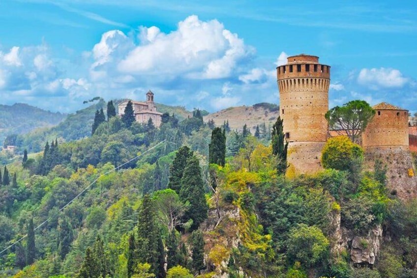 Ein historischer Stein-Turm und eine Festung auf einer Anhöhe, umgeben von üppiger Vegetation und anderen Gebäuden. 1