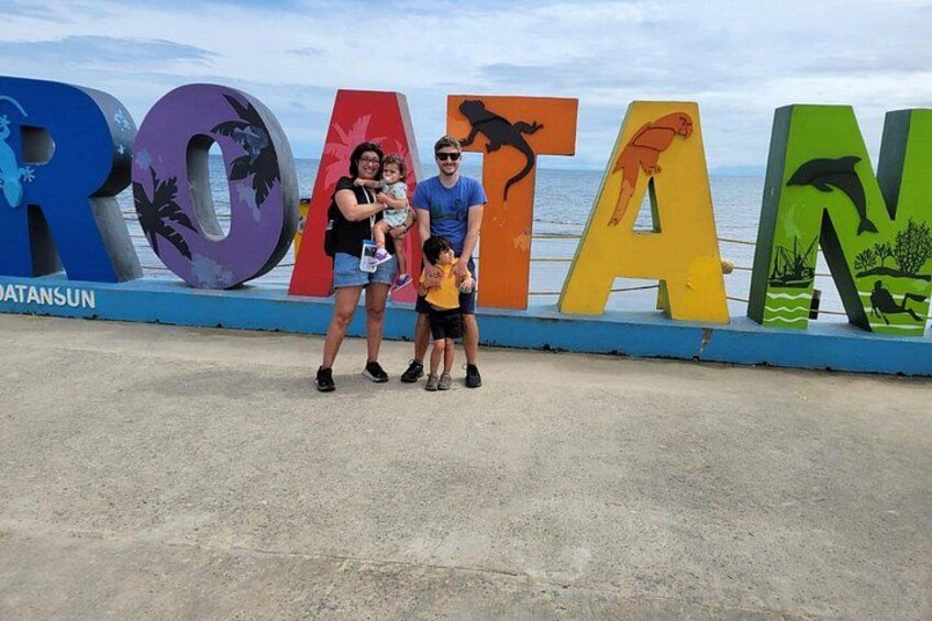 Una familia parada frente a letras grandes y coloridas que deletrean 'ROTA', con una playa y el océano al fondo. 3
