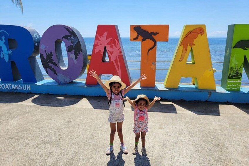 Dos niños de pie frente a letras coloridas que deletrean 'ROATAN', con una playa y el océano al fondo. 5