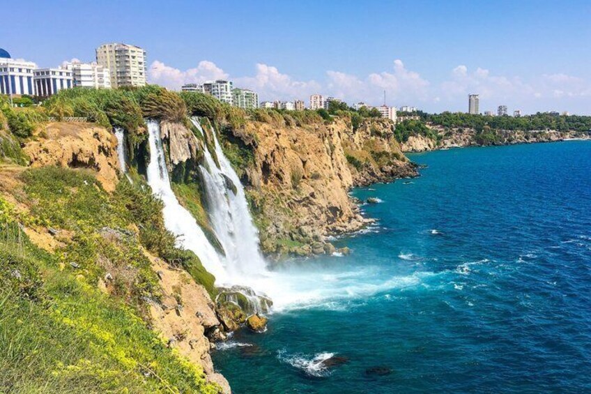 A coastal waterfall, clear blue sea, and urban buildings in the background. 2