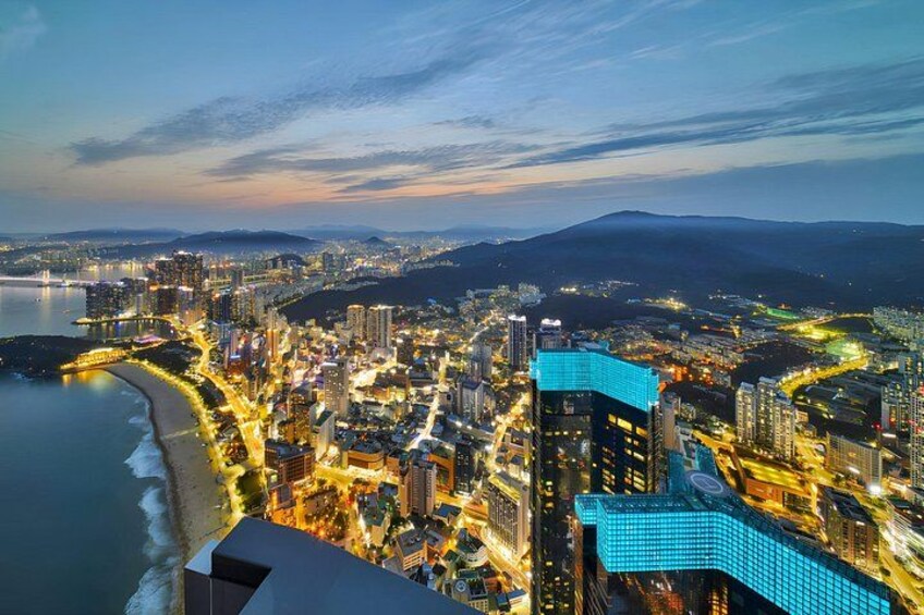 Un paisaje urbano costero al anochecer con edificios iluminados, una playa y una montaña al fondo. 1