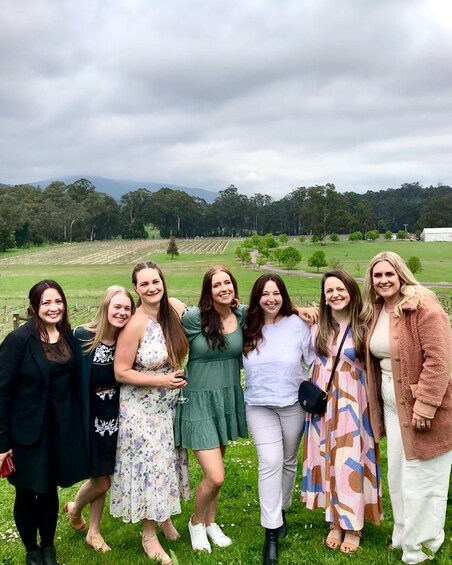 A group of people standing on grass with a vineyard and trees in the background. 3