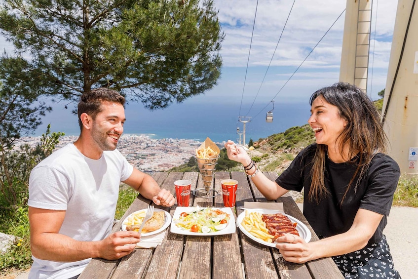 Two people sitting at a wooden table with food and drinks, overlooking a coastal landscape with a cable car in the background. 1