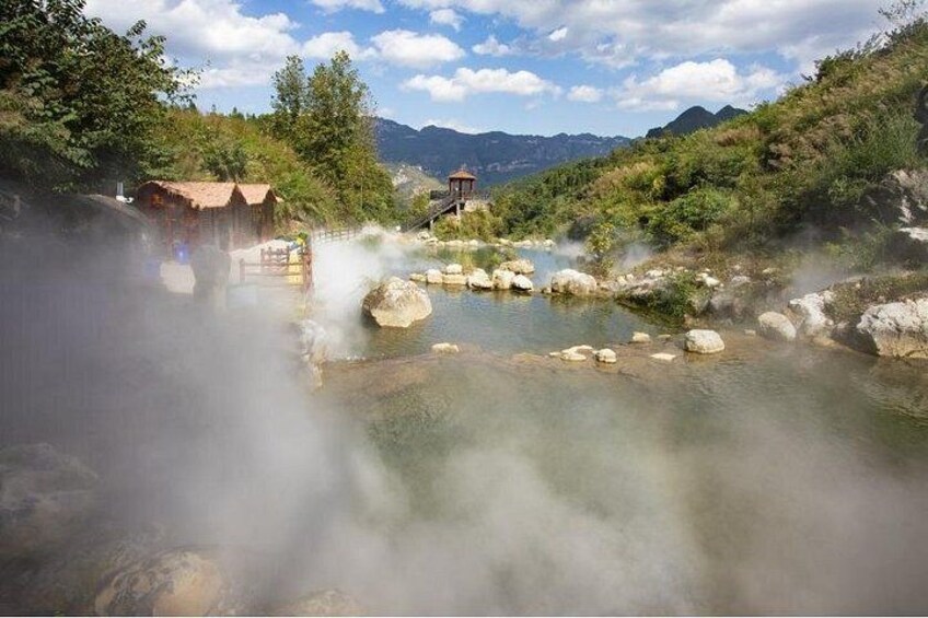 A hot spring with steam rising, surrounded by greenery and a wooden structure. 5