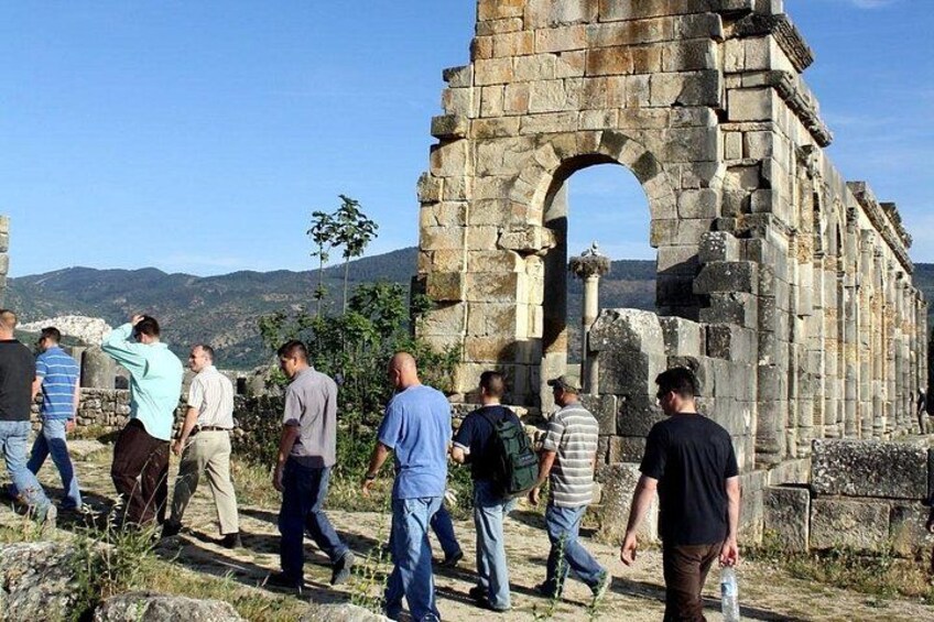 Eine Gruppe von Menschen spaziert in der Nähe alter Steinstrukturen mit einer Bergkulisse. 1
