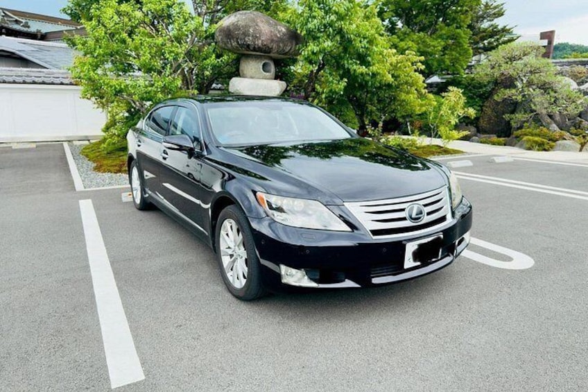 Un coche Lexus negro estacionado en una carretera con líneas blancas, rodeado de vegetación y una estructura de piedra. 3