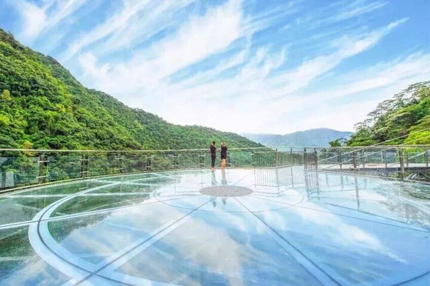 A glass-bottomed walkway with a person standing on it, surrounded by lush green hills and a clear sky. 2