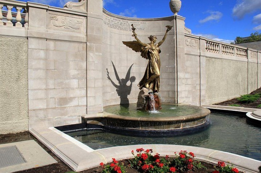 Una estatua de bronce de una mujer sosteniendo una antorcha, ubicada en una fuente y rodeada por un muro de piedra con inscripciones. 5