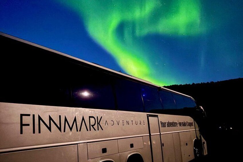 A bus with 'FINNMARK ADVENTURE' written on it, parked under a night sky with the Northern Lights. 1