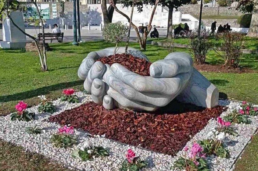 A sculpture of hands holding a heart in a park with flowers and a bench in the background. 2
