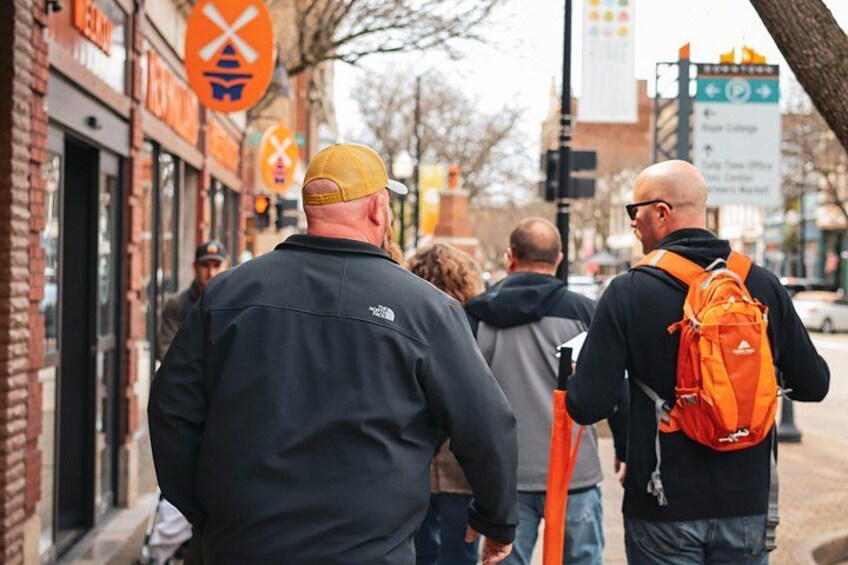 Eine Gruppe von Menschen geht auf einer Straße entlang, gesäumt von Backsteingebäuden und orangefarbenen Schildern. 2