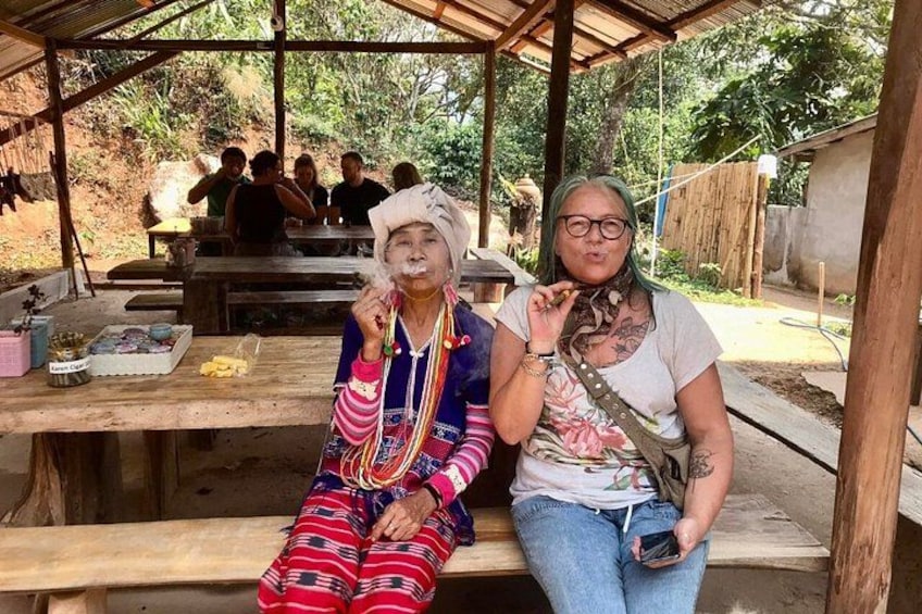 Two people sitting on a wooden bench outdoors, with a group of people in the background. 5