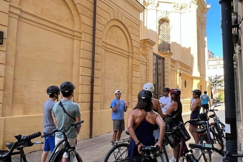 Een groep mensen op fietsen verzameld voor een historisch gebouw met gebogen ramen en een poort. 5