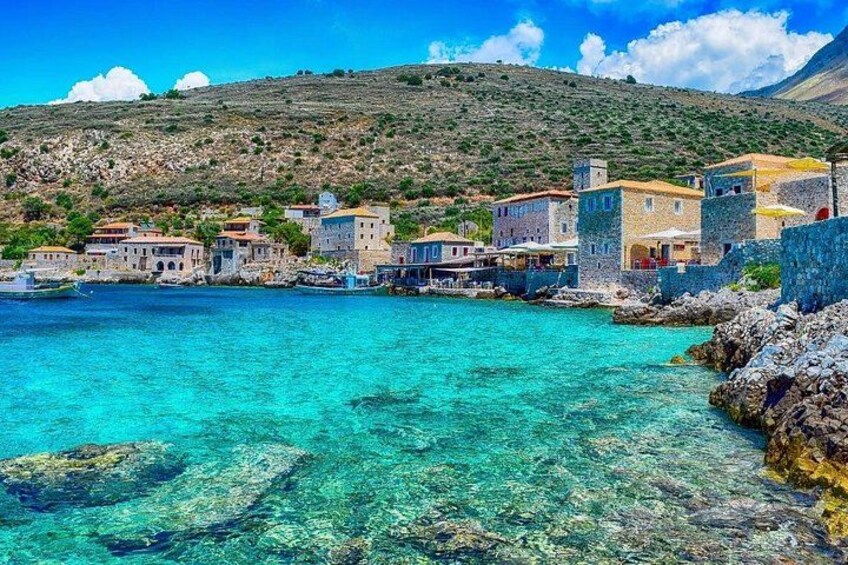 A coastal village with clear turquoise waters, stone buildings, and a hilly backdrop. 1
