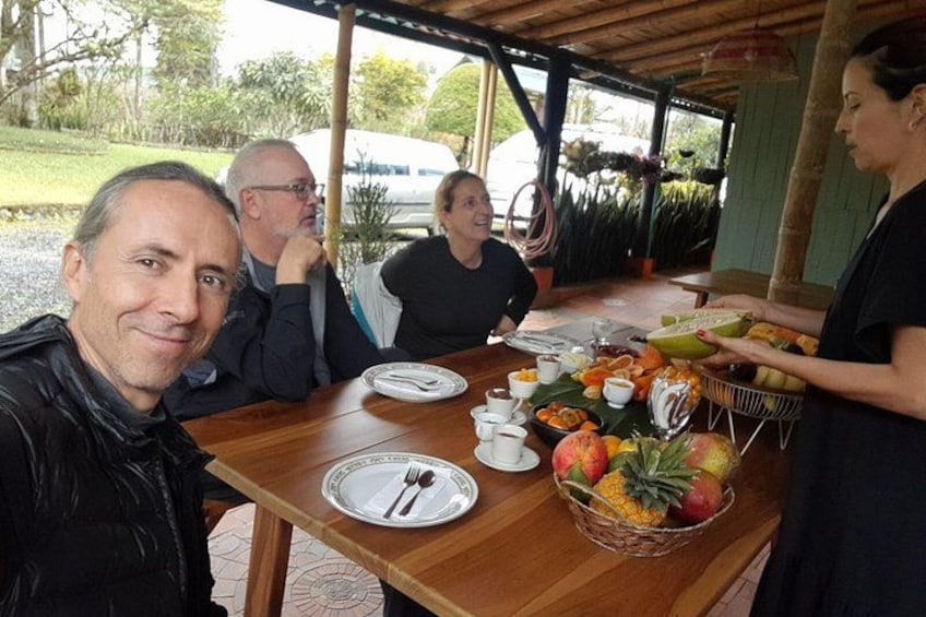 En gruppe mennesker spiser ved et udendørs bord med forskellig frugt og drikkevarer. 1
