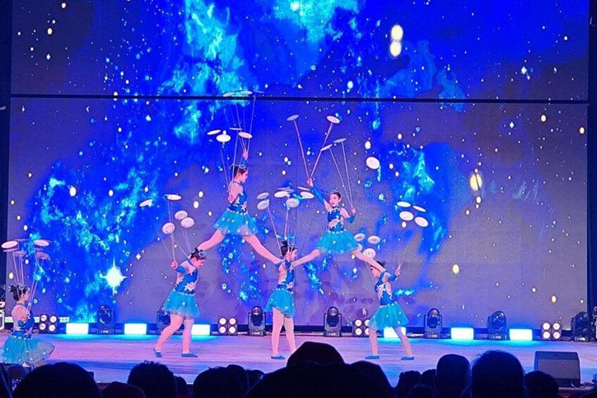A stage performance with dancers in blue costumes, a blue-themed backdrop, and stage lighting. 2