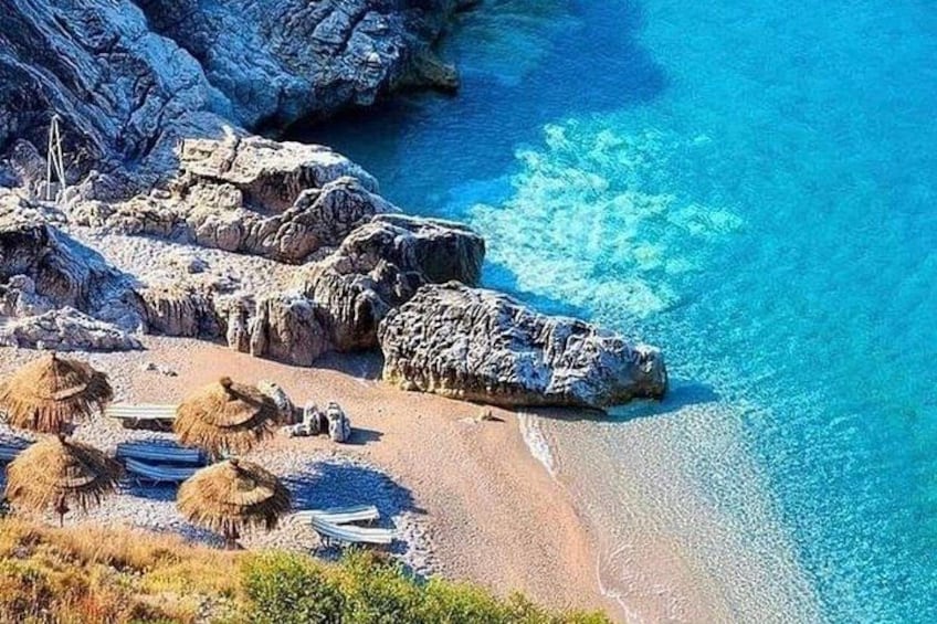 A rocky coastline with clear blue water, a sandy beach, and thatched-roof huts. 1
