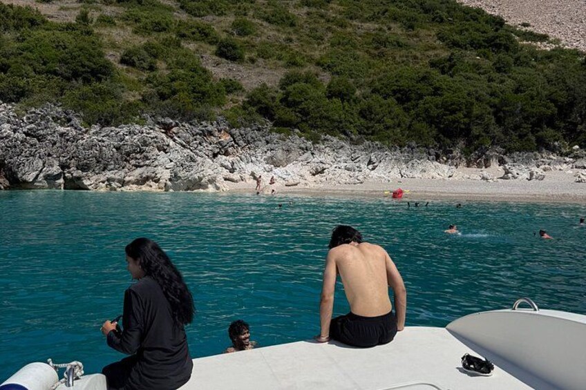 A boat with people on a rocky shore, clear blue water, and a beach with people swimming. 5