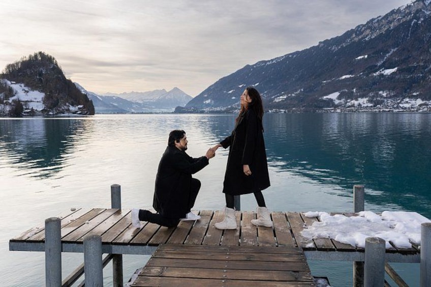 Ein Mann macht einer Frau an einem Holzsteg am See einen Heiratsantrag, im Hintergrund Berge. 1