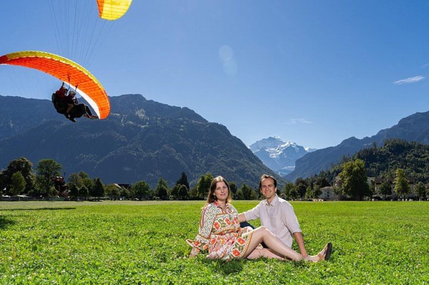 Ein Paar sitzt auf einer Wiese, im Hintergrund Berge und ein Paraglider am Himmel. 3
