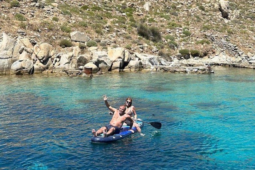 Två personer på en blå flotte i klart vatten, med en klippig kustlinje i bakgrunden. 3