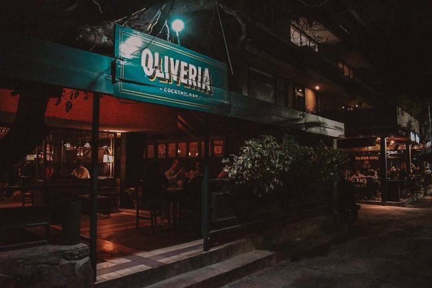 Aftenstemning fra en bar ved navn 'Oliveria' med udendørs siddepladser og en neonskilte. 5
