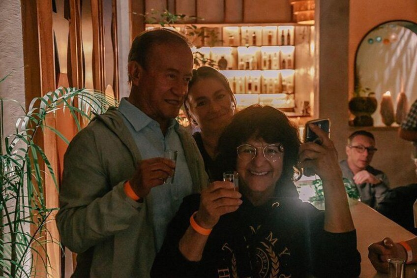 En person tager et selfie med en smartphone, en bar med flasker i baggrunden, og en anden person holder et glas med drik. 4