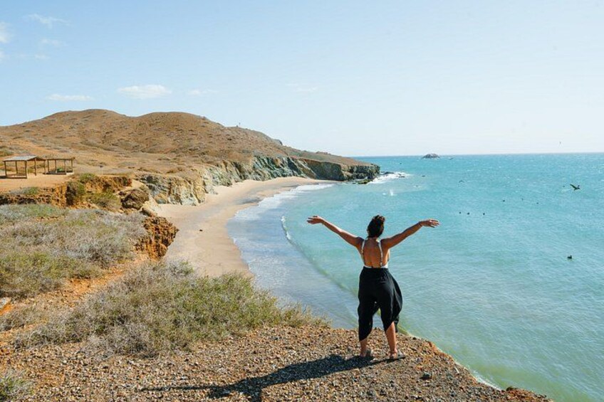 A person standing on a rocky beach with arms outstretched, overlooking a calm sea and a hilly landscape. 2