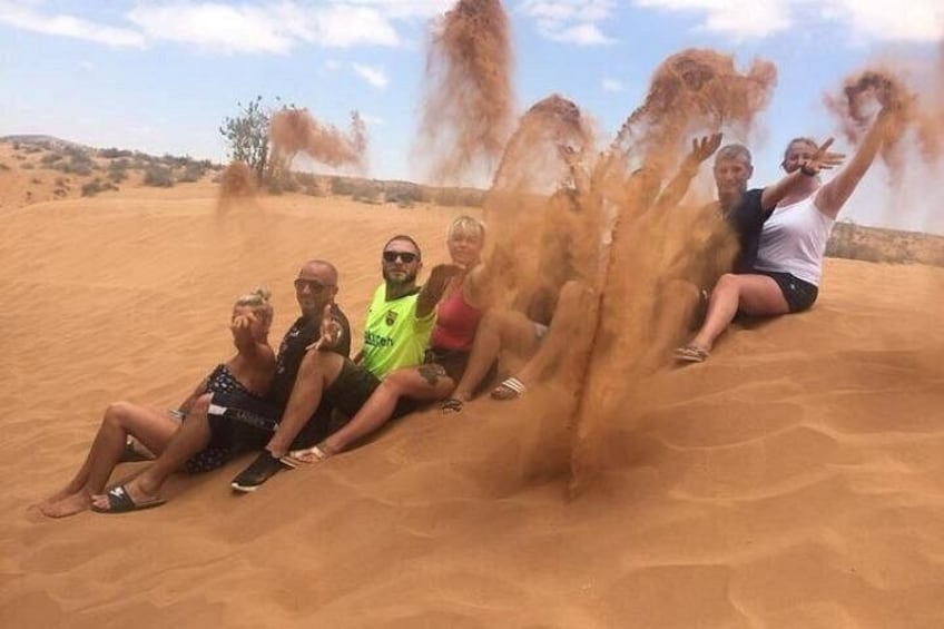 En grupp människor sitter på en sandstrand och sand virvlar runt omkring dem. 1
