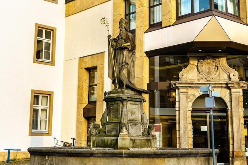 A statue of a robed figure on a pedestal, a building with a yellow facade, and a street lamp. 2