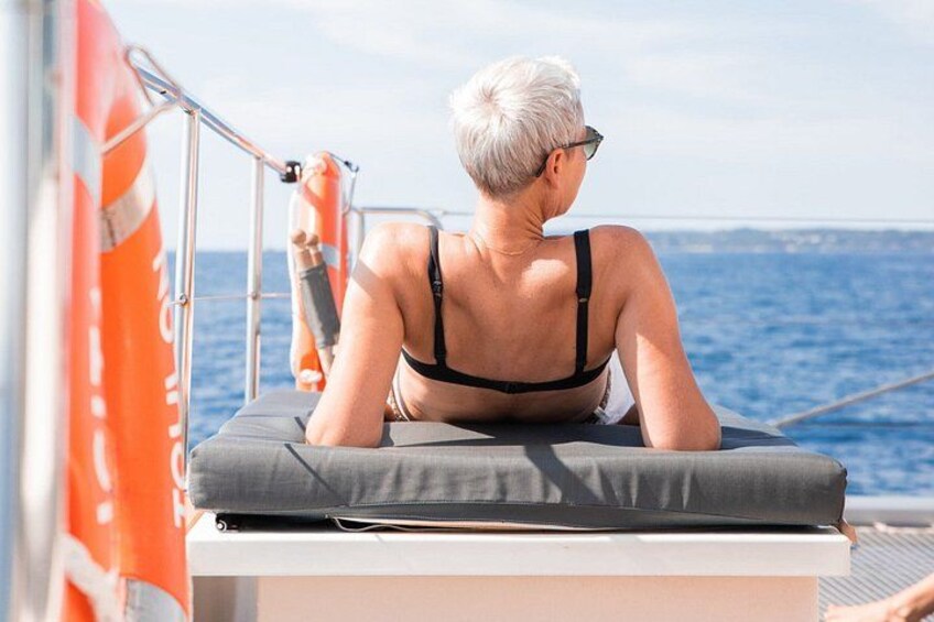 A person sitting on a boat deck, wearing a black swimsuit, with a lifebuoy and a flag in the background. 1