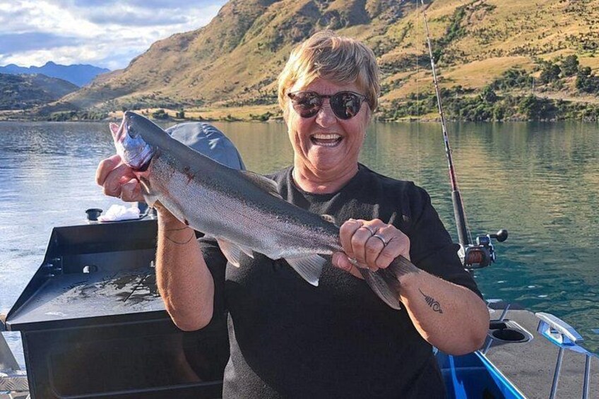 A person holding a fish on a boat with a lake and mountains in the background. 5