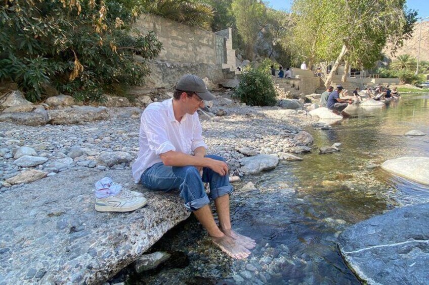 A person sitting on a rocky ledge by a clear stream, with others in the background. 3