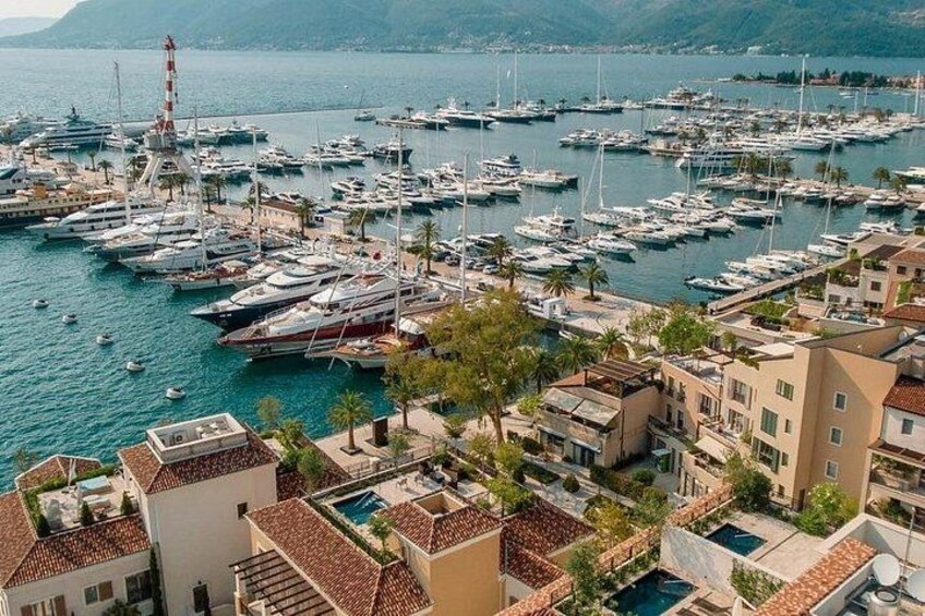 A marina with numerous boats docked, buildings with red-tiled roofs, and a mountainous landscape in the background. 2