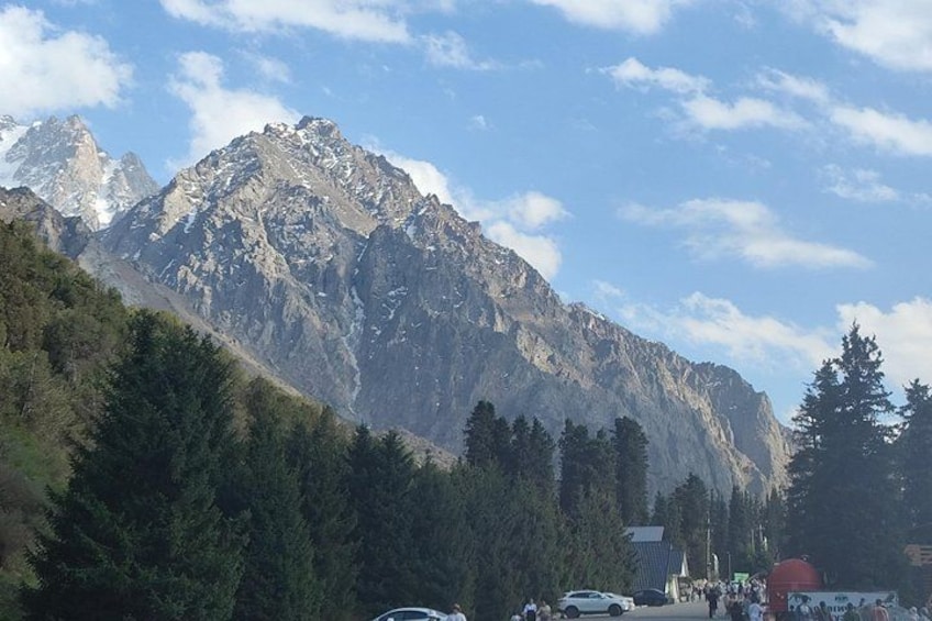 A mountain range with a clear sky, a forested area in the foreground, and a few vehicles and people. 1