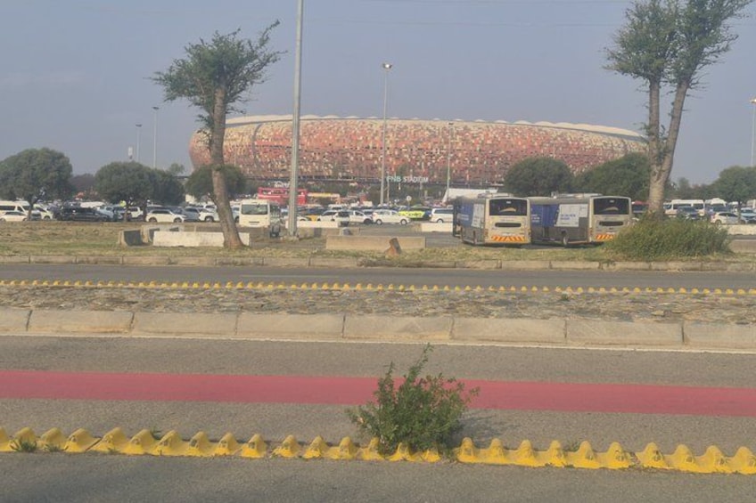 Un estadio con buses y coches estacionados, una carretera con una zona pintada de rojo y amarillo, y árboles. 2