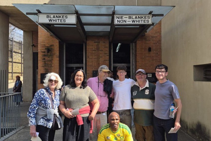 Un grupo de personas de pie frente a un edificio con letreros que dicen 'BLANKES WHITES' y 'NIE BLANKES NON WHITES'. 3