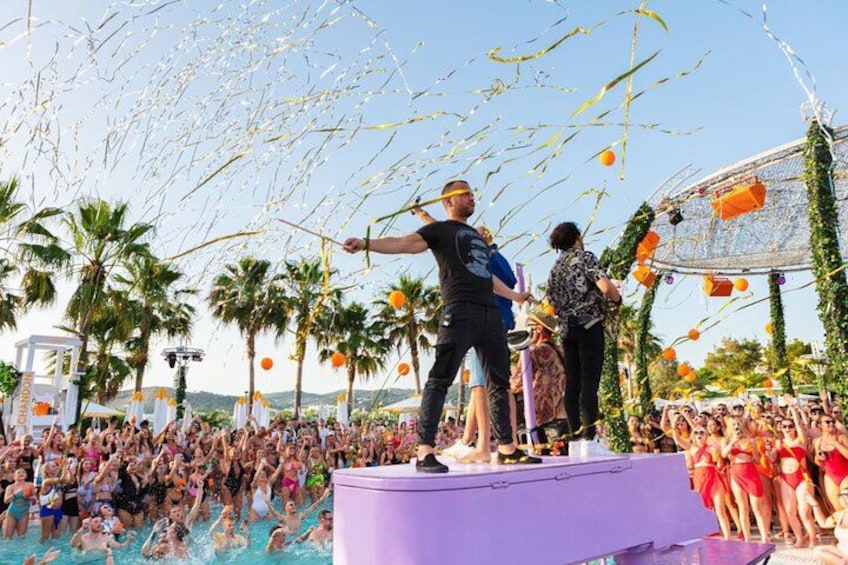 A stage with people performing, a crowd in the background, palm trees, and a clear sky. 3