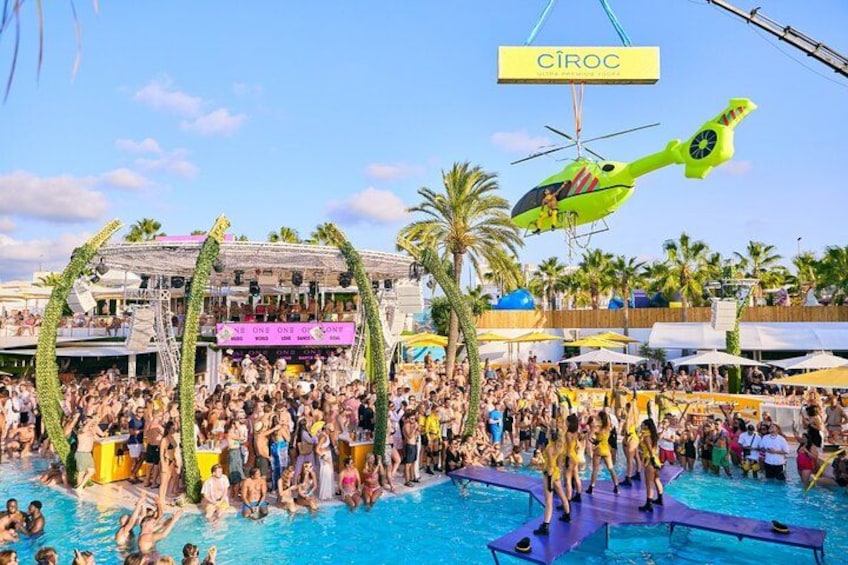 A crowded outdoor swimming pool with a helicopter and a sign that reads 'CIROC' in the background. 2