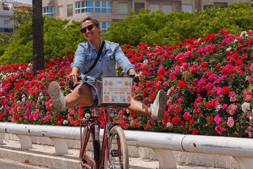 Una persona sentada en una bicicleta con un letrero en el frente, rodeada por un jardín con flores rojas y rosadas. 1