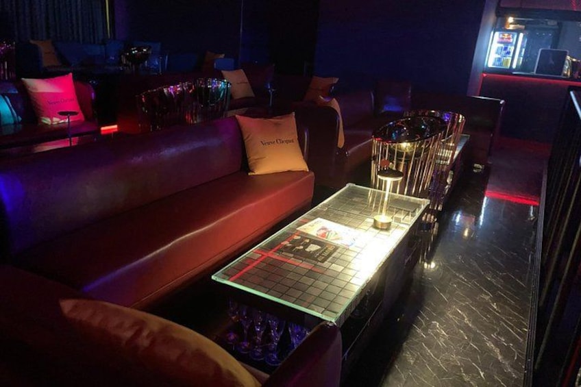 A dimly lit lounge area with a red leather couch, a glass-top coffee table with a grid pattern, and a bar with a neon sign. 3