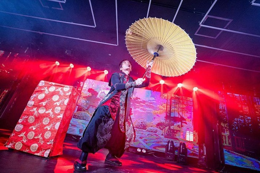 A person in traditional attire holding an umbrella on a stage with a red-lit background and a large screen displaying text. 1