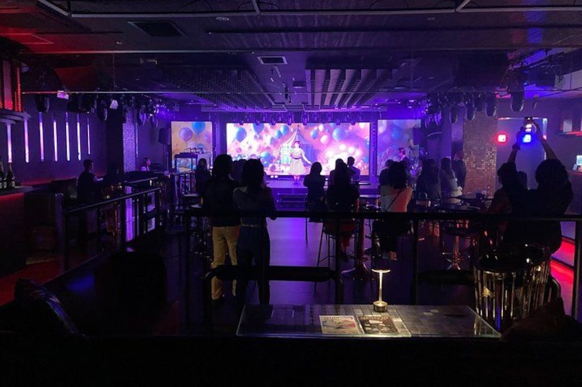 A dimly lit bar with a stage, people seated at tables, and a DJ booth. 5