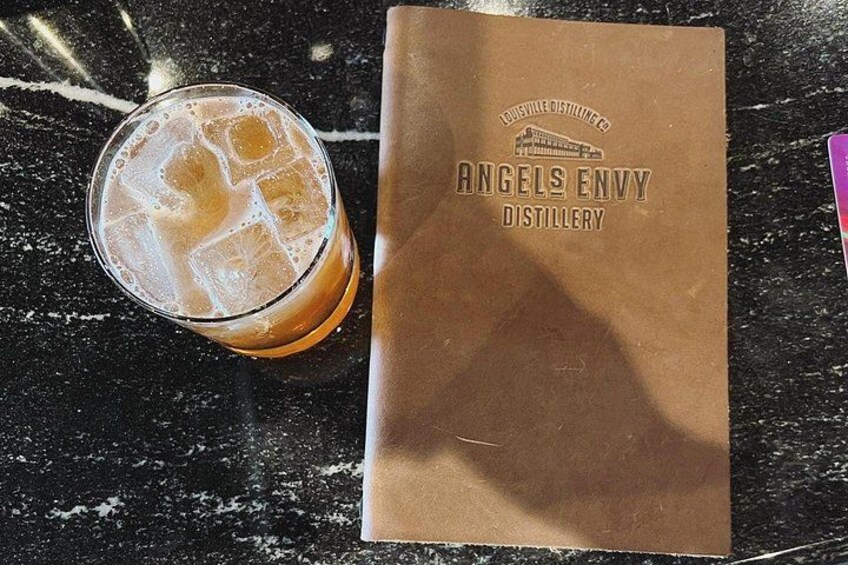 A glass of iced beverage and a brown paper bag with 'Angels Envy Distillery' printed on it. 2