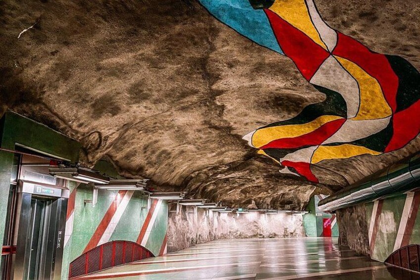 Une station de métro souterraine dont le plafond est orné d’une fresque colorée et les murs de motifs rayés. 3